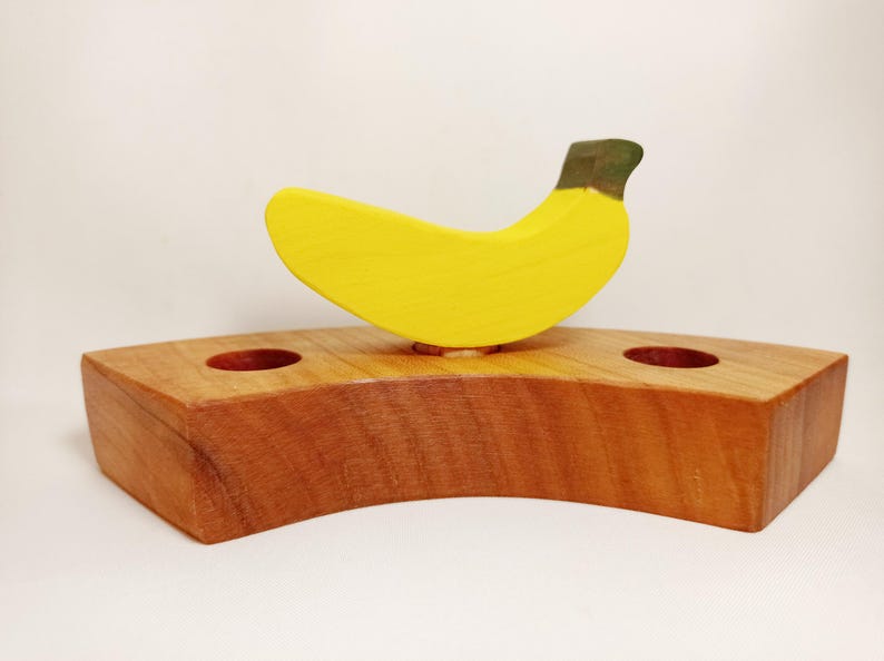 May include: A wooden banana shaped toy with a yellow body and a green stem sits on a wooden base with two holes. The base is brown and has a curved shape.