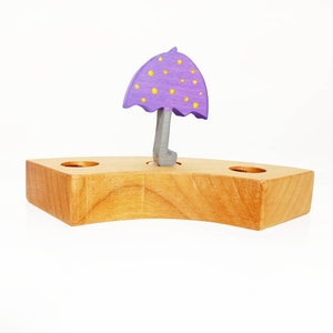 Può includere: Un giocattolo di legno con un ombrello viola con puntini gialli sopra una base di legno curva.