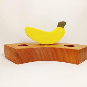 May include: A wooden banana holder with two holes for toothbrushes. The banana is yellow with a green stem and is made of wood.