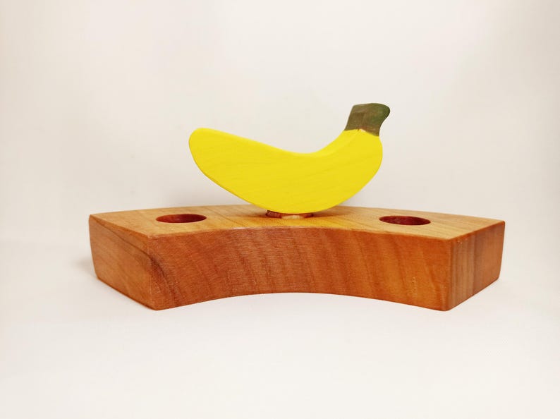 May include: A wooden banana holder with two holes for toothbrushes. The banana is yellow with a green stem and is made of wood.