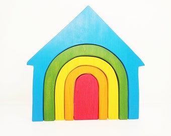 May include: A rainbow-colored wooden house toy with a blue roof and a red door. The house is made of five interlocking arches, each a different color of the rainbow.