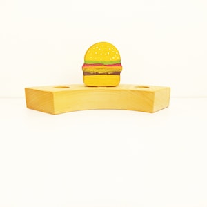 May include: A wooden toy hamburger on a wooden stand with two holes. The hamburger is yellow with brown and red details.