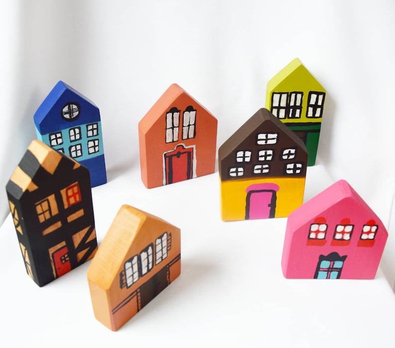 May include: A selection of seven brightly painted wooden house ornaments. Each house is a different colour, such as blue, orange, yellow, pink, and green, with white window and door details. The houses vary in size and design.