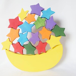 Può includere: Una luna gialla a forma di mezzaluna con una pila di stelle di legno colorate in cima. Le stelle sono in un motivo arcobaleno, con stelle rosse, arancioni, gialle, verdi, blu e viola.