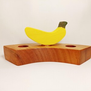 May include: A wooden banana-shaped candle holder with two holes for candles. The holder is made of natural wood and has a yellow banana painted on top.