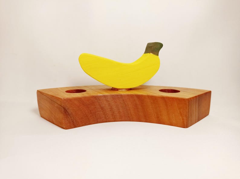 May include: A wooden banana-shaped candle holder with two holes for candles. The holder is made of natural wood and has a yellow banana painted on top.