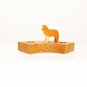 May include: A wooden toy with a brown and white wolf figure standing on a wooden base with three holes. The base is shaped like a crescent moon.