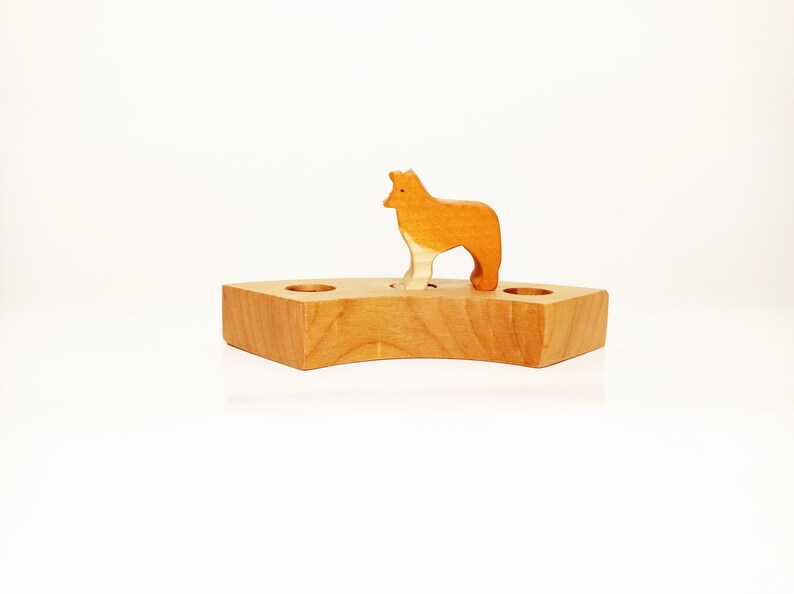 May include: A wooden toy with a brown and white wolf figure standing on a wooden base with three holes. The base is shaped like a crescent moon.