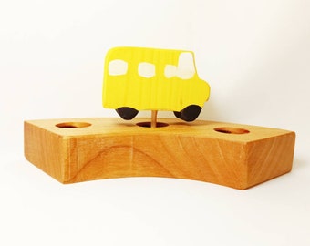 May include: A wooden toy school bus, painted yellow with white windows and black wheels, sits atop a natural wood base. The base has three holes and a curved edge, creating a playful and decorative item.