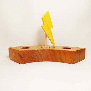 May include: A wooden candle holder featuring a yellow lightning bolt. The holder is made of light brown wood and has two circular openings. The lightning bolt is mounted on a small wooden stand.