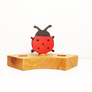 以下が含まれることがあります： 木製のてんとう虫のキャンドルホルダー。てんとう虫は赤色で黒い斑点と黒い頭があります。キャンドルホルダーのベースは、2つの円形のキャンドル用の開口部がある、明るい茶色の幾何学的な形状です。背景は白です。