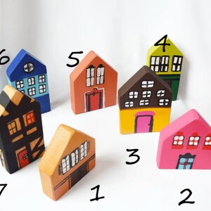 May include: A selection of seven hand-painted wooden houses in various colours. Each house has a unique design, with different window arrangements and door styles. The houses are painted in shades of orange, pink, yellow, green, blue, and black, creating a charming, whimsical display.
