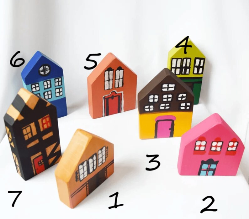 May include: A selection of seven hand-painted wooden houses in various colours. Each house has a unique design, with different window arrangements and door styles. The houses are painted in shades of orange, pink, yellow, green, blue, and black, creating a charming, whimsical display.