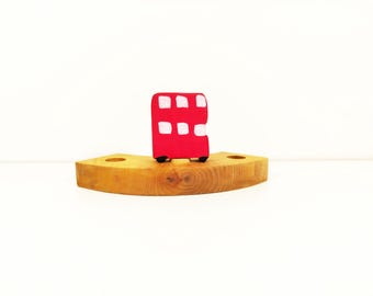 May include: A red and white toy double-decker bus sits on a wooden base. The bus has a simple design with white windows and black wheels.