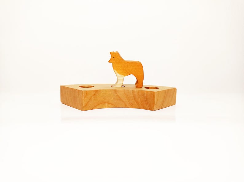 May include: A wooden toy with a small wooden animal figure standing on top. The animal is a light brown color and has a white chest. The base of the toy is a light brown color and has three holes in it.