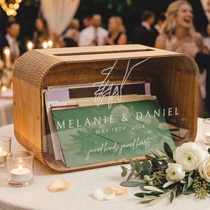 May include: A rectangular wooden card box with a clear acrylic front panel featuring a white line drawing of joined hands, the text "MELANIE & DANIEL", "MAY 18TH, 2028", and "joined hands, joined hearts" printed in white over a green envelope.