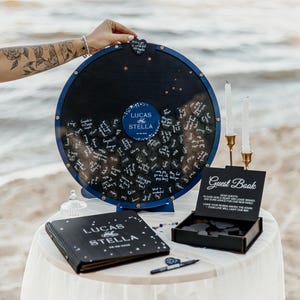 May include: A navy blue wedding guest book alternative with a circular design. The center features the names "Lucas and Stella" in white script. A matching guest book and a "Guest Book" sign are also present.