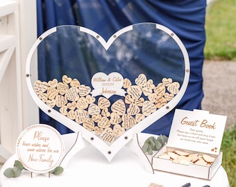 May include: A white heart-shaped box with a clear lid filled with wooden heart-shaped cards for guests to sign at a wedding. The box is on a white table with a sign that says "Please leave wishes and some advice for our New Family".