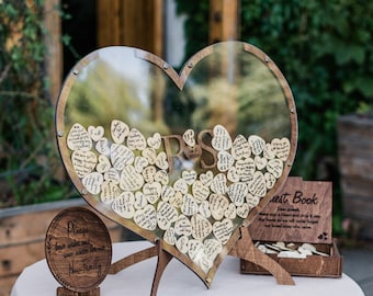 May include: A wooden heart-shaped guest book with a clear acrylic cover filled with small wooden hearts with handwritten messages. The guest book is on a table with a sign that says "Please leave wishes and love advice for our new family".