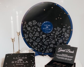 May include: A wedding guest book set featuring a large, round, black sign-in board with heart-shaped tags. A matching guest book and a box of heart-shaped tags are also present. The board has the names "LUCAS STELLA" and the date.