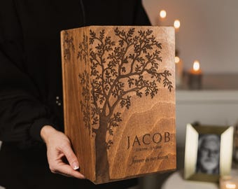 Può includere: Una scatola di legno con un disegno di albero inciso. Il nome "JACOB" è visualizzato, insieme a date e la frase "forever in our hearts". La scatola è tenuta da una persona che indossa una camicia nera.