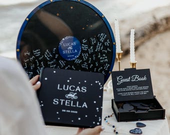 May include: A wedding guest book set with a black and blue theme. The set includes a large circular sign-in board, a guest book, and a box of heart-shaped sign-in cards. The sign-in board and guest book feature the names "LUCAS STELLA".