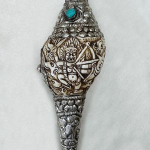 May include: Ornate, silver-toned conch shell with intricate carvings and a turquoise stone accent. The shell features detailed relief carvings of figures and patterns, with a cone-shaped base and a decorative top. The text "Himalayan Healers" is visible.