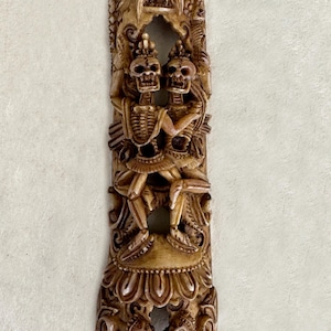 Può includere: Una scultura in legno intagliato raffigurante due scheletri che si abbracciano, con dettagli intricati e una finitura marrone chiaro. La scultura ha una forma verticale e allungata con intagli ornati di figure e motivi floreali.