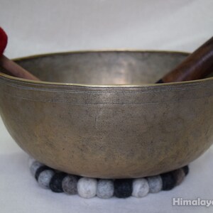 May include: A large, antique-looking singing bowl with a wooden striker and a red felt mallet. The bowl is resting on a gray and black felt cushion.