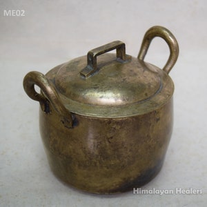 Op de afbeelding: Een kleine, antieke koperen pot met een deksel en twee handvatten. De pot heeft een versleten, verweerde afwerking en wordt waarschijnlijk gebruikt om te koken of te serveren.