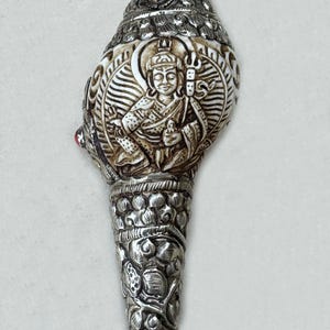 May include: Ornate silver-colored conch shell with intricate carvings and inlaid gemstones. Features a central relief of a seated figure and floral patterns. The shell has a conical shape and is likely a decorative or ceremonial item.