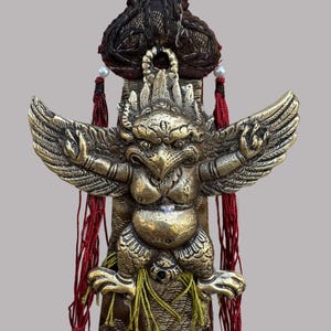 May include: A detailed brass sculpture of a mythical creature with outstretched wings, intricate detailing, and a textured surface. The sculpture is mounted on a brown fabric base with red and green decorative elements.