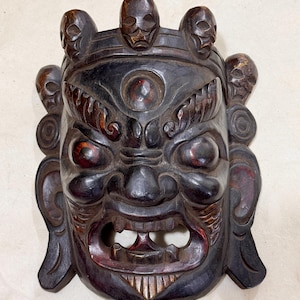 May include: A dark brown carved wooden mask with a fearsome expression. The mask has red accents, a wide open mouth, and skull details. It is likely of cultural or spiritual significance, possibly from a Himalayan tradition.