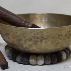 May include: A brass singing bowl with a hammered finish, resting on a grey and white felt cushion. A wooden striker with a red felt tip is included.