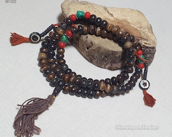 Puede incluir: Un collar de cuentas de mala de madera marrón con detalles rojos y turquesa. El collar tiene un borla en un extremo y un pequeño amuleto de ojo en el otro. El collar se muestra sobre una pieza de madera.