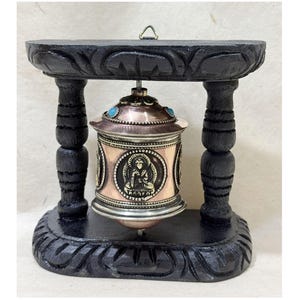 May include: A small, copper-colored prayer wheel with a silver trim and turquoise accents. The wheel is mounted on a black wooden stand with intricate carvings.