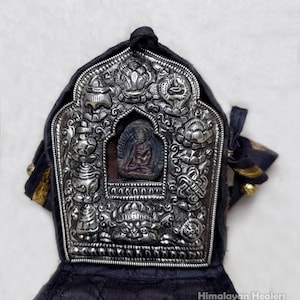 May include: A silver-colored religious pendant with intricate carvings and a central Buddha figure. The pendant is set against a dark fabric background, with gold accents and the text "Himalayan Healers" at the bottom.