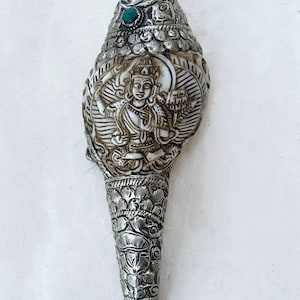 Puede incluir: Concha de plata ornamentada con una figura de deidad tallada y detalles turquesa. La concha presenta intrincados diseños florales y geométricos, con una base cónica y una parte superior redondeada. El texto "SA01" es visible en la parte superior.