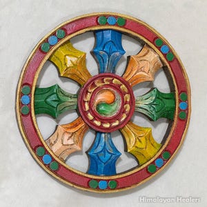Puede incluir: Una colorida rueda del Dharma de madera con un anillo exterior rojo y detalles dorados. La rueda presenta ocho radios en varios colores, incluyendo azul, verde, amarillo y naranja. El centro tiene un diseño circular con un remolino verde y naranja.