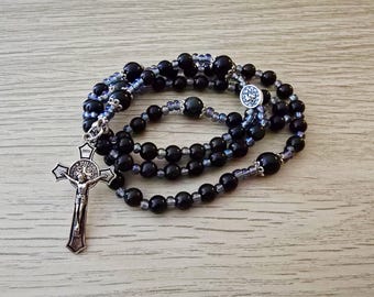 Puede incluir: Un rosario negro con un crucifijo plateado y un emblema circular. Las cuentas son una mezcla de negro y azul-gris translúcido. El rosario está sobre una superficie de madera clara.
