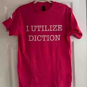 May include: A bright pink t-shirt with the words "I UTILIZE DICTION" printed in white block letters. The shirt is hanging on a white hanger against a white door.