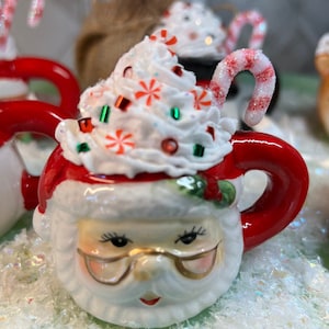 May include: A white ceramic Santa Claus mug with a red handle and a white whipped cream topping with red and green candy sprinkles. The mug has a candy cane sticking out of the whipped cream.