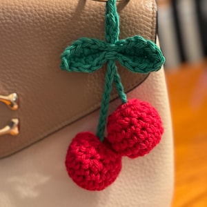 May include: A handmade crochet cherry keychain with two red cherries, green leaves, and a green stem. The keychain is attached to a silver ring. The background is a beige purse.