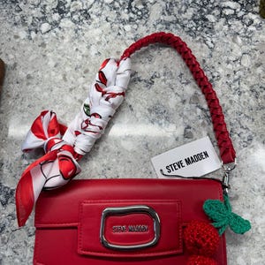 May include: A red handbag with a braided handle and a decorative scarf. The bag features a silver buckle and the brand name "STEVE MADDEN". Adorned with crocheted cherries and a green leaf.