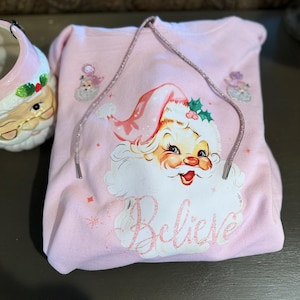 May include: A light pink hooded sweatshirt featuring a Santa Claus graphic with the word "Believe" in a cursive font. The Santa graphic has a red hat and a white beard. The sweatshirt has a drawstring and small snowflake and angel graphics.
