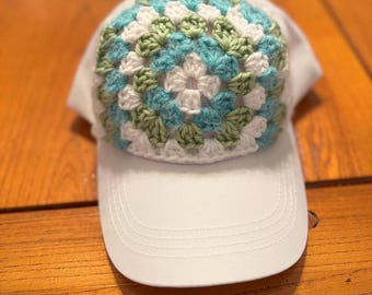 May include: White baseball cap with a crocheted design in shades of blue, green, and white. The crocheted pattern is circular and covers the top of the cap, adding a unique, handmade touch to the accessory.