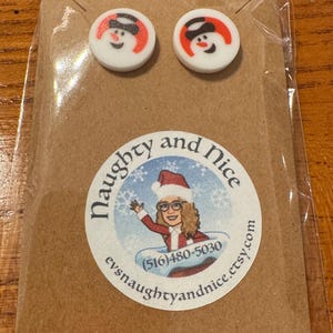 May include: A pair of snowman stud earrings on a brown card. The earrings are white with a red and black snowman design. The card has a circular sticker with the text "Naughty and Nice" and a cartoon image of a woman in a Santa hat.