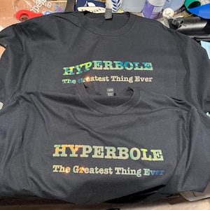Puede incluir: Dos camisetas negras con la palabra "HYPERBOLE" en letras iridiscentes multicolores. Debajo, la frase "The Greatest Thing Ever" está impresa en una fuente más pequeña.