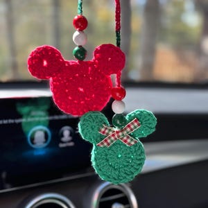 May include: Two crochet Mickey Mouse head car charms. One is red, the other is green with a plaid bow. Both have beaded hanging strings with red, white, and green beads. The charms are hanging in front of a car's dashboard.