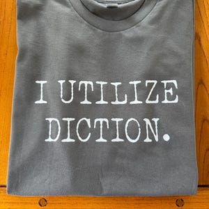 May include: A gray t-shirt with the text "I UTILIZE DICTION." printed in white. The shirt is folded neatly on a wooden surface. The t-shirt has a crew neck.